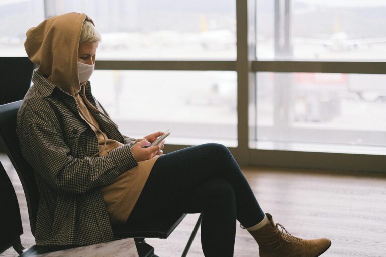 A woman in a hoodie and face mask uses a smartphone while waiting at an airport terminal.