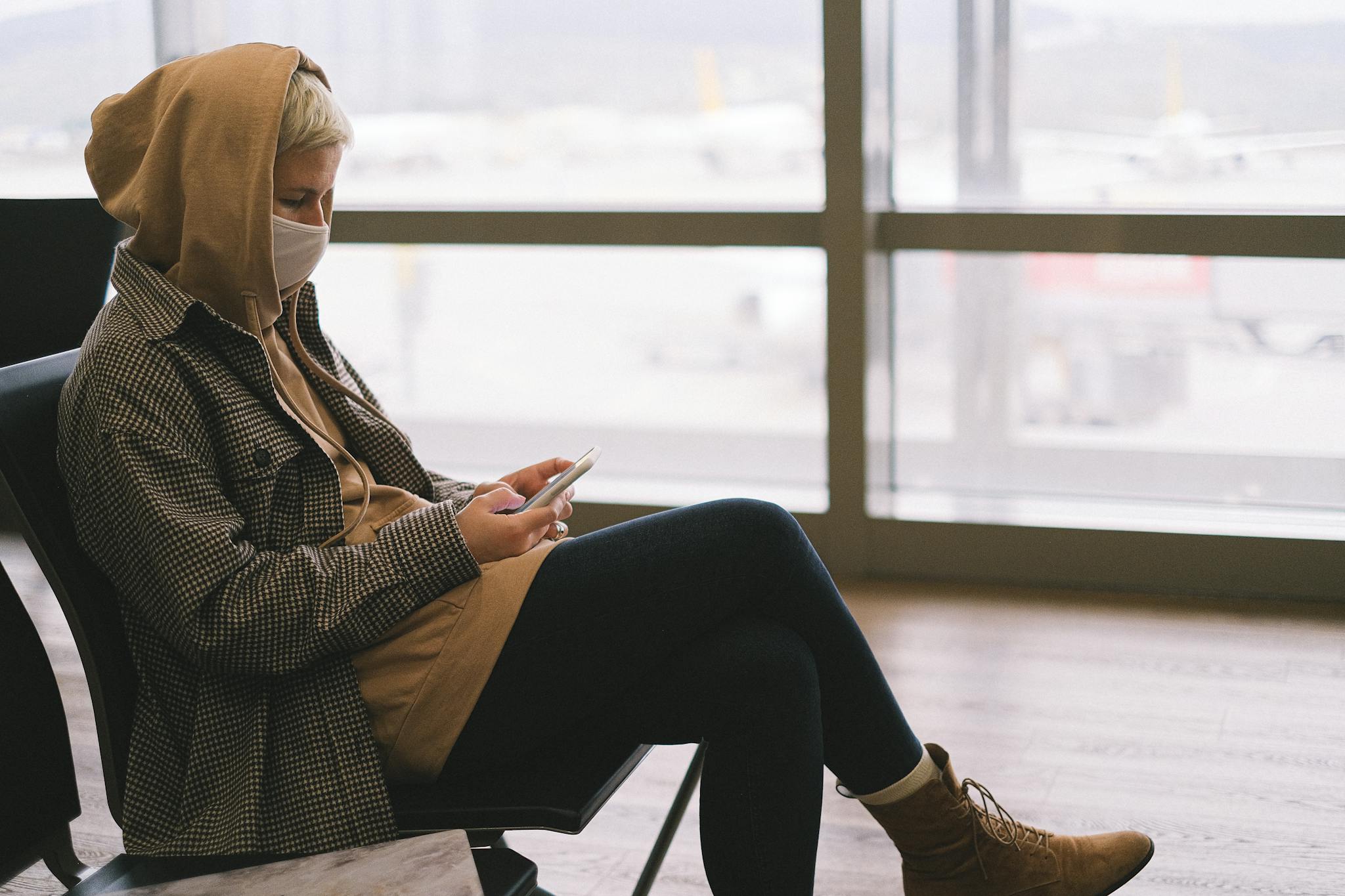 A woman in a hoodie and face mask uses a smartphone while waiting at an airport terminal.